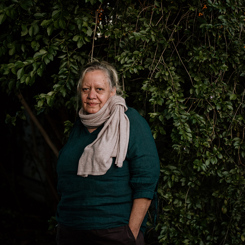 Debbie Carmody stands in front of foliage. She is looking directly into the camera lens and her left hand is in her pocket. She is wearing a forest green long sleeved shirt and beige scarf.