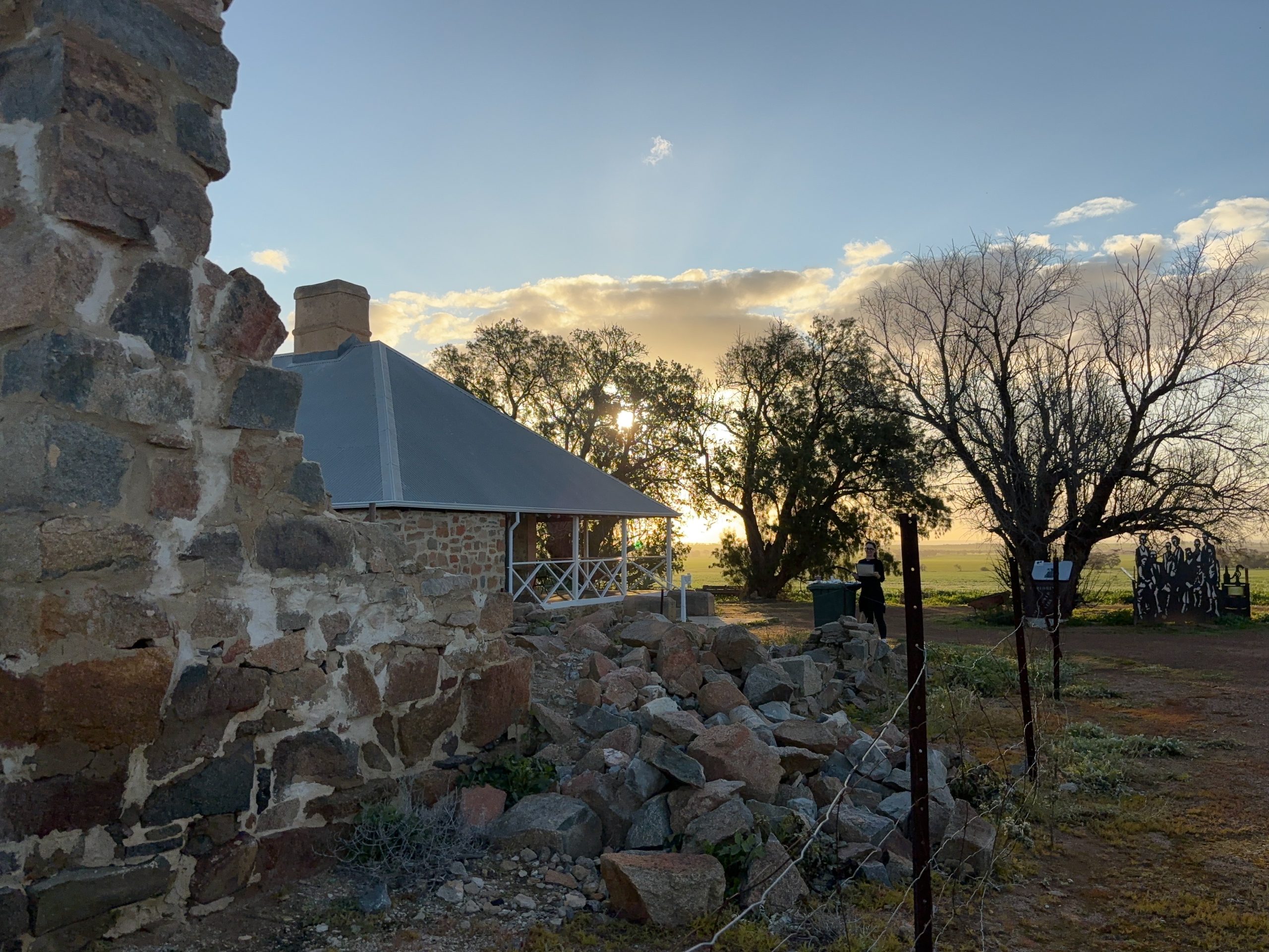 Naomie stands outside the MacPherson Homestead which is surrounded by a fence and a leafy tree. The sun is setting in the background.